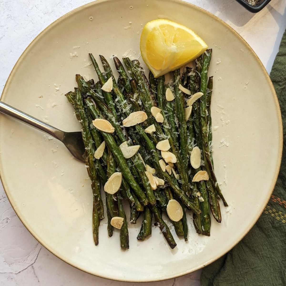 Green beans on a white plate with almonds scattered on top and a lemon wedge at the side.