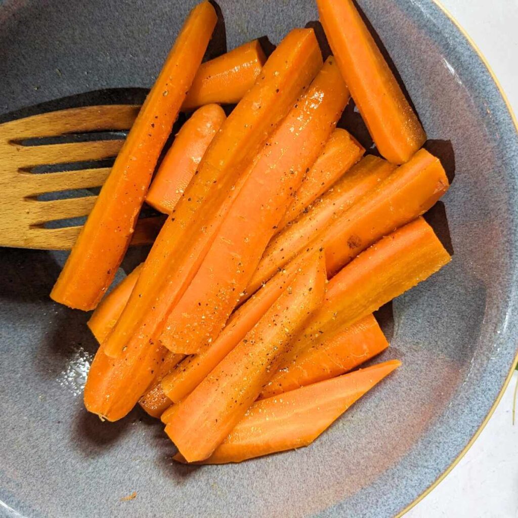 Carrot sticks in a bowl with a wooden spoon which is mixing the seasoning onto the carrots.