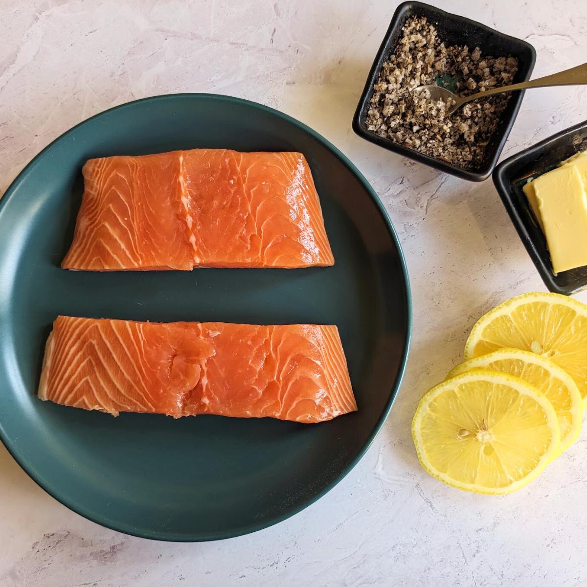Ingredients for making salmon in an air fryer with two salmon fillets on a plate, slices of lemon, butter, salt and pepper