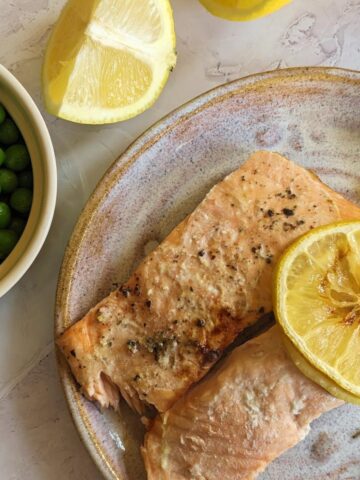 Two salmon fillets on a plate with a slice of lemon on top and a bowl of peas at the side.