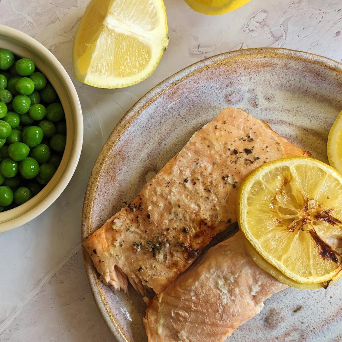 Two salmon fillets on a plate with lemon slices on top. A bowl of peas sits at the side.