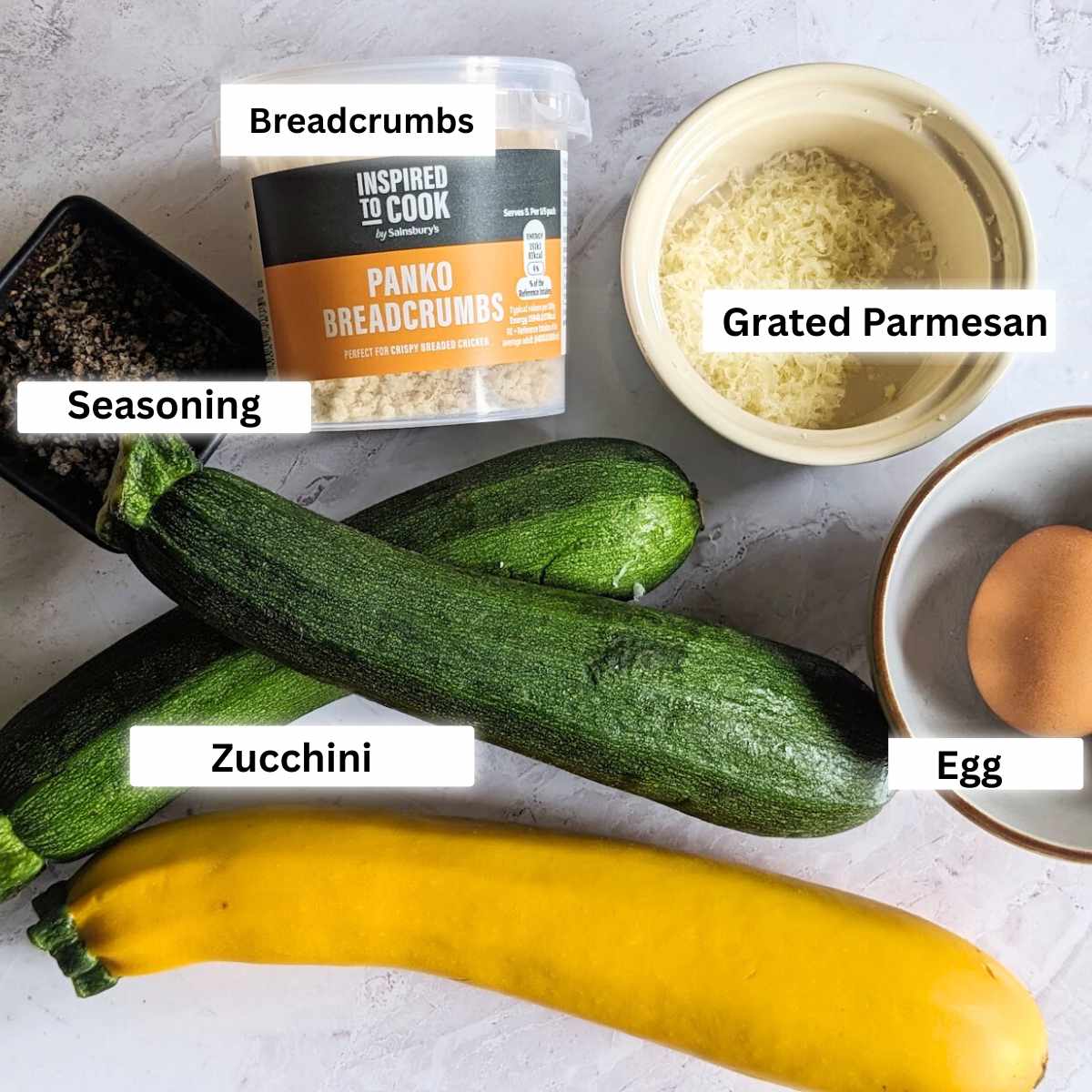 Ingredients for making zucchini fries on a board, including zucchini, breadcrumbs, seasoning, grated parmesan and an egg.