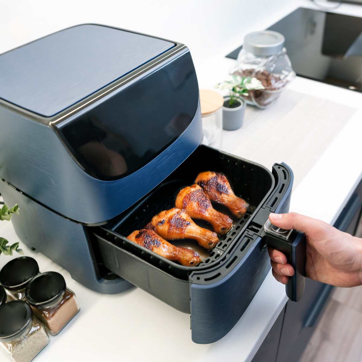 Cooking chicken drumsticks in an air fryer basket with a hand opening the basket.